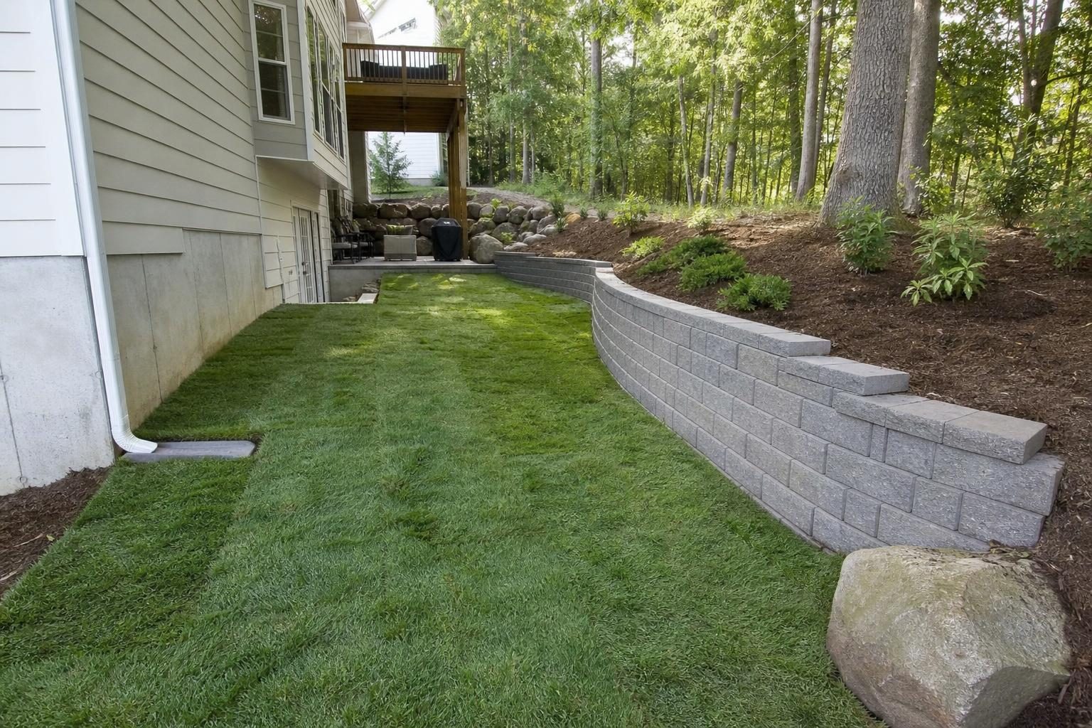 A low block retaining wall terraces the steep part of the slope, while the gentler area beside it was regraded so water drains away from the house.