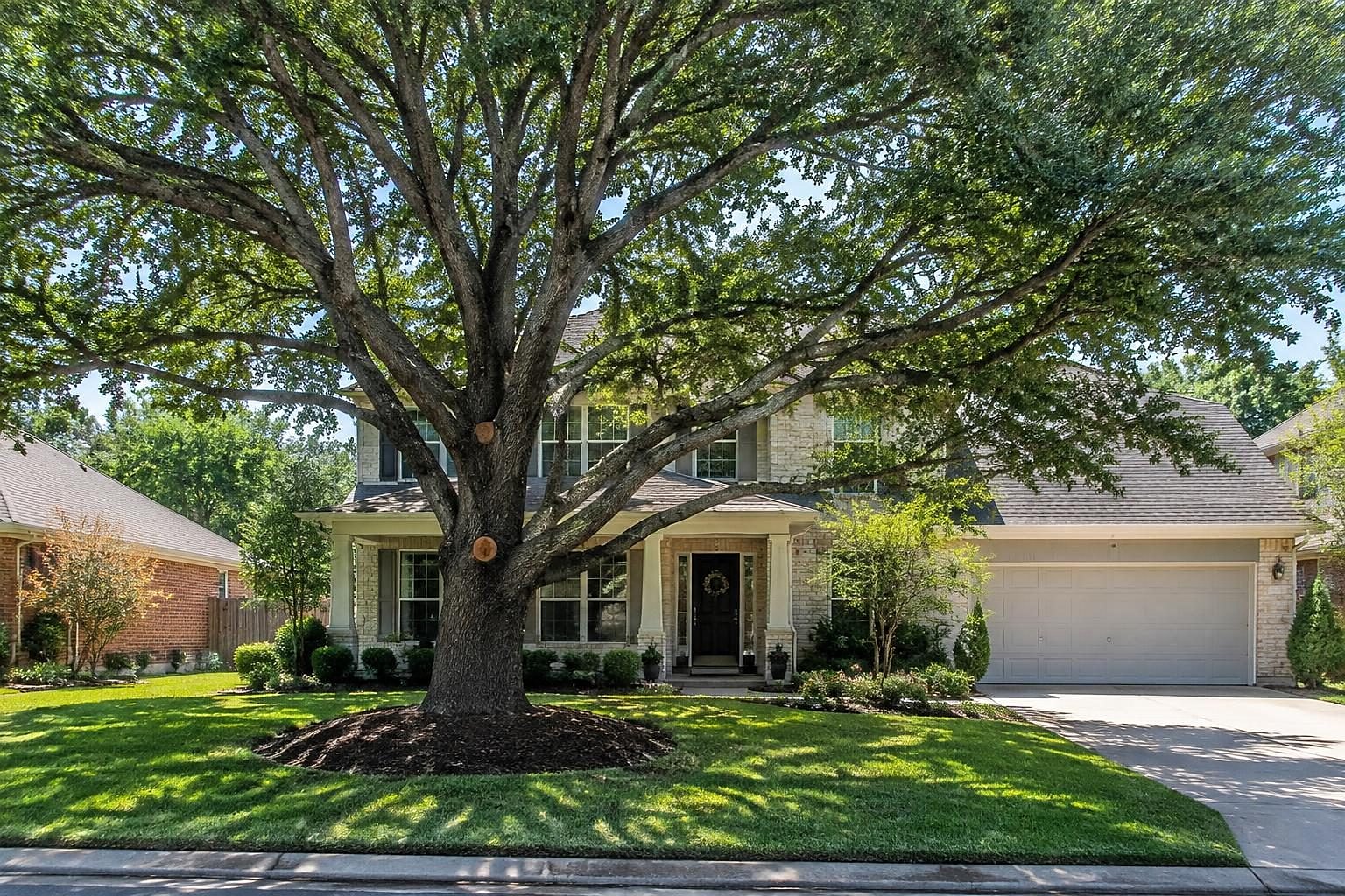 A large, well-shaped mature oak tree with a mulch ring shading the front yard of a two-story suburban house